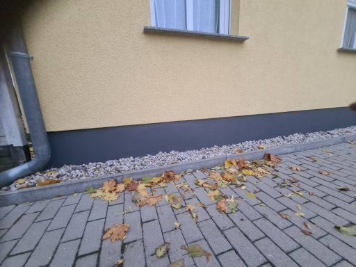 Grau gestrichene Wand mit Kies und herabgefallenen Blättern auf dem Boden.
