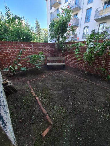 Kleiner Garten mit einer Bank, umgeben von grünen Pflanzen und Ziegelmauer.